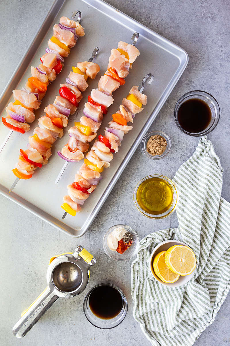 Ingredients for chicken skewers including diced chicken breasts, sliced red, orange, and yellow bell peppers, chopped yellow onion, and marinade ingredients like olive oil, soy sauce, balsamic vinegar, brown sugar, lemon juice, and seasonings.